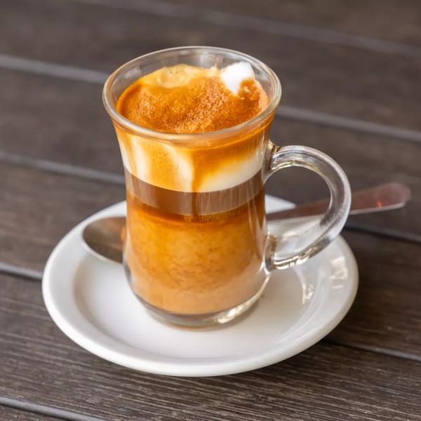 A layered coffee drink with foam in a clear mug on a white saucer with a spoon.