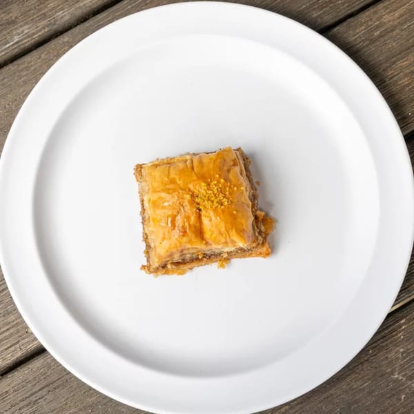 A single square piece of baklava, drizzled with syrup and sprinkled with nuts, on a white plate.