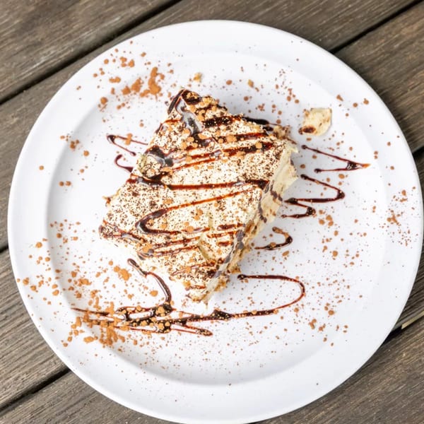 A slice of tiramisu with chocolate drizzle and cocoa powder on a white plate.