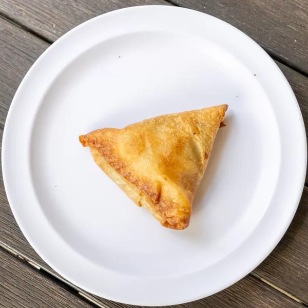 A single golden-brown samosa rests on a white plate, centered on a rustic wooden table.